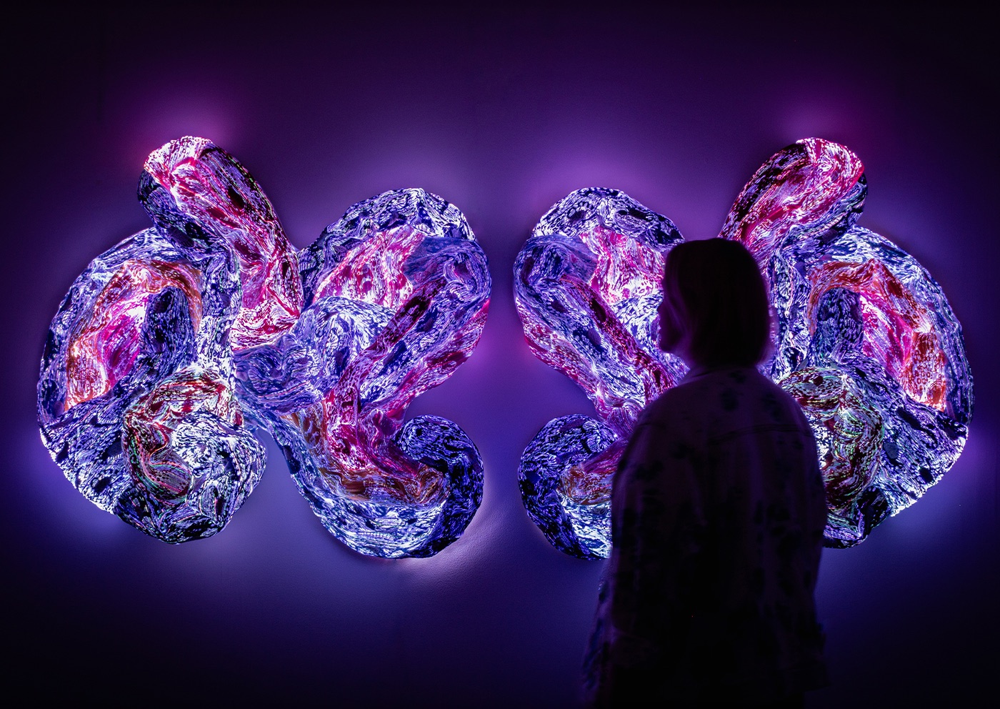 Självlysande textilverk i lila och rosa ljus på en mörk vägg, med en besökare i siluett framför, på Andetag Museum i Stockholm