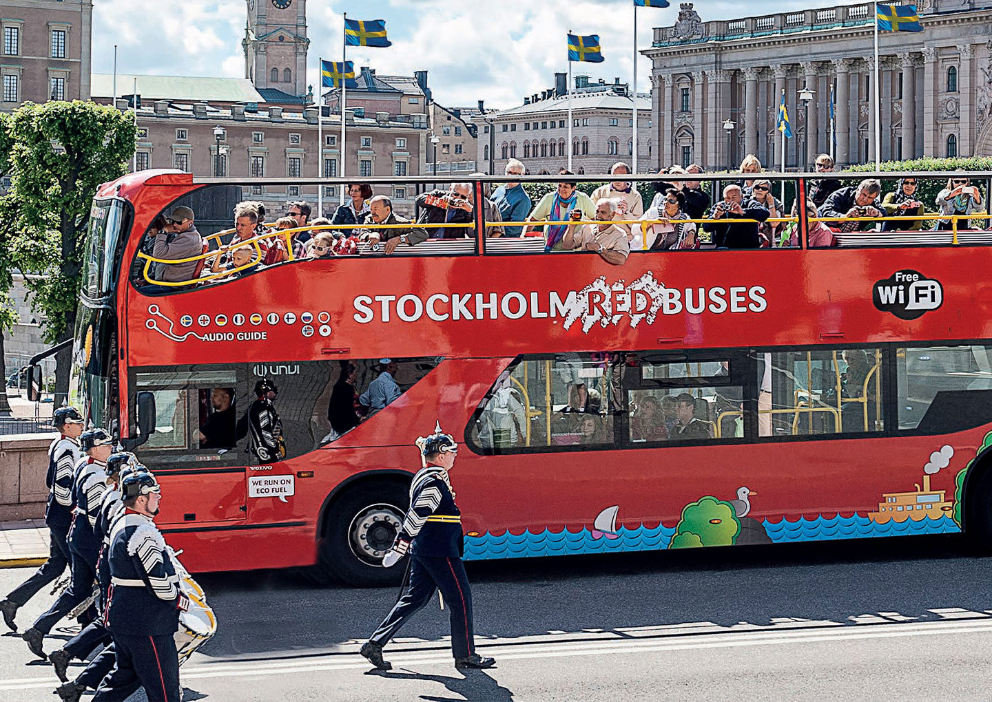 Röd dubbeldäckad sightseeingbuss med öppen övervåning kör förbi Riksdagshuset medan en militärorkester marscherar på gatan