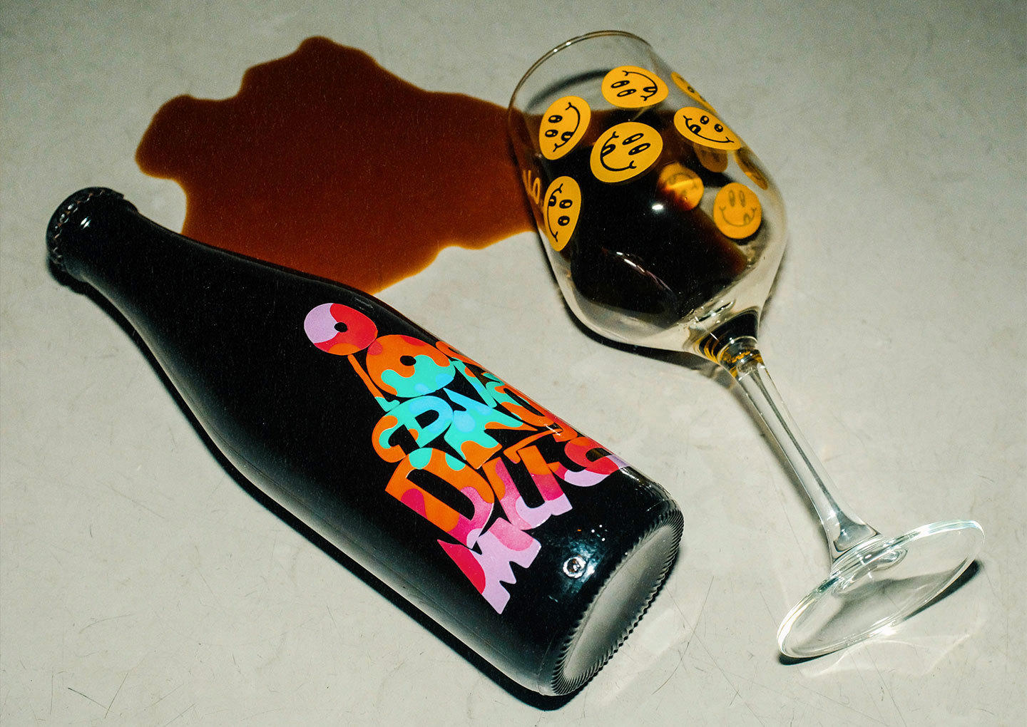 Mörk ölflaska från Omnipollo med färgglad etikett bredvid ett vinglas med smileymönster och utspilld öl