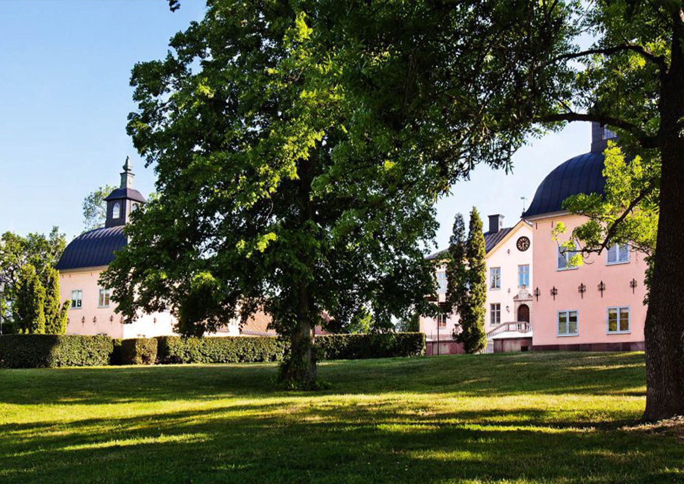 Hesselby Slotts rosa slottsbyggnader bakom grönskande träd och gräsmatta en solig sommardag