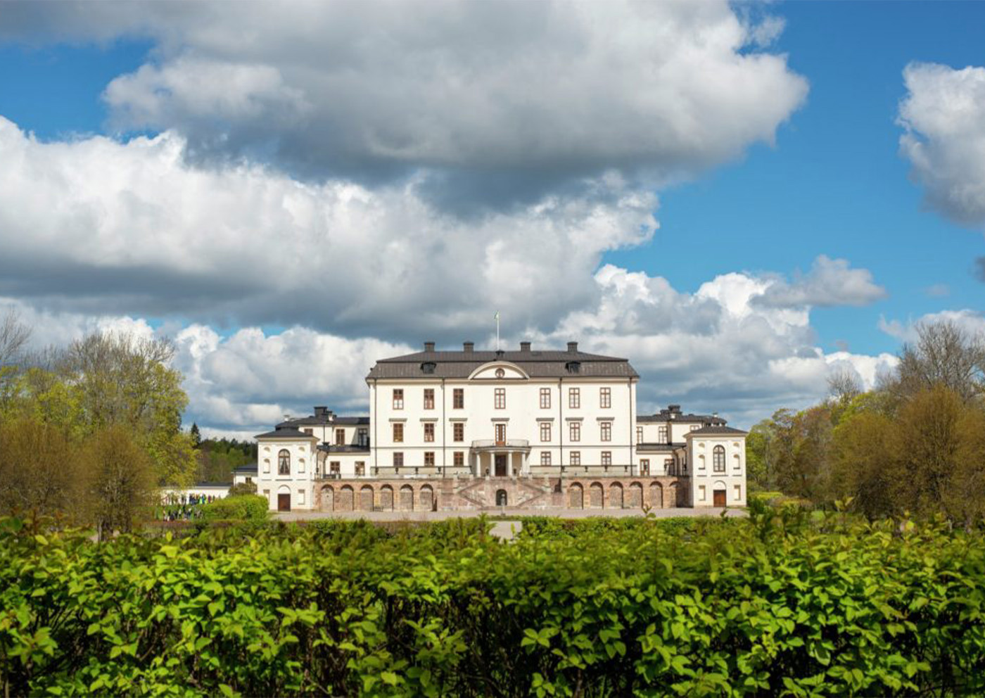 Rosersbergs slott med vit huvudbyggnad och flyglar bakom grön häck en solig sommardag